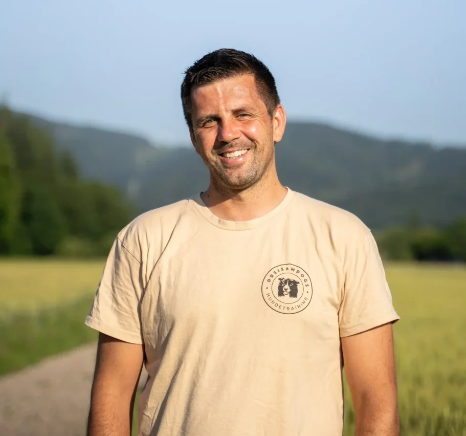 Lächelnder Mann in T-Shirt steht vor einer Landschaft mit Bergen und Wiesen.